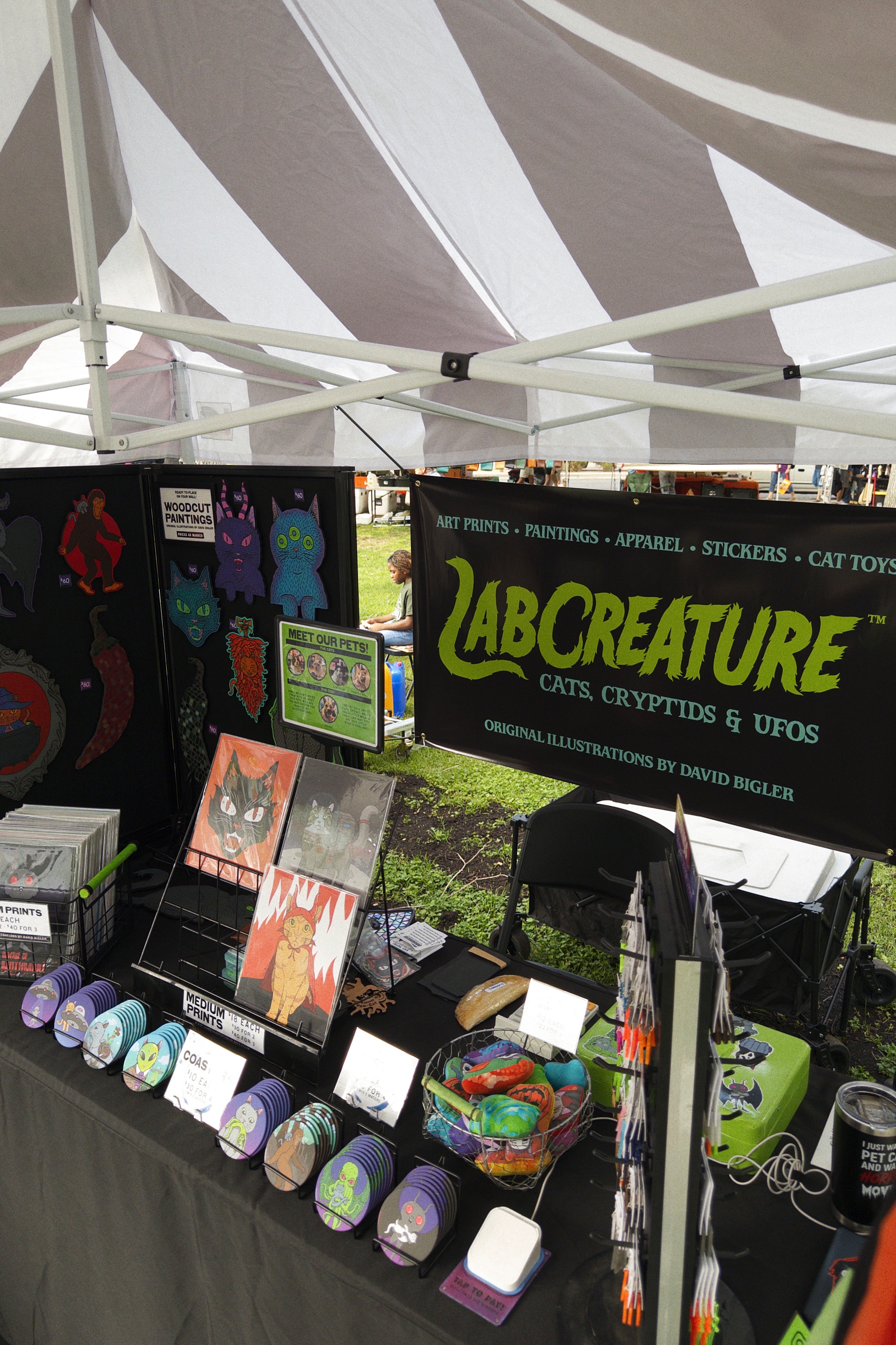 A vendor booth under a black and white striped tent with a black banner that says art prints, paintings, apparel, stickers, and cat toys, underneath there’s a hand lettered green logo for labcreature, original illustrations by David Bigler. In the front are assorted coasters, prints, cat toys on a black tablecloth. 