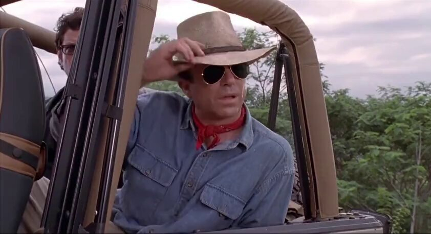 Screenshot from Jurassic Park of Alan Grant as played by the late great Sam Neill who is holding the brim of a cowboy hat, wearing sunglasses, and a red neckerchief. He is sitting in an open jeep, with greenery in the background. This is when he is looking at dinosaurs at the beginning of the film. 