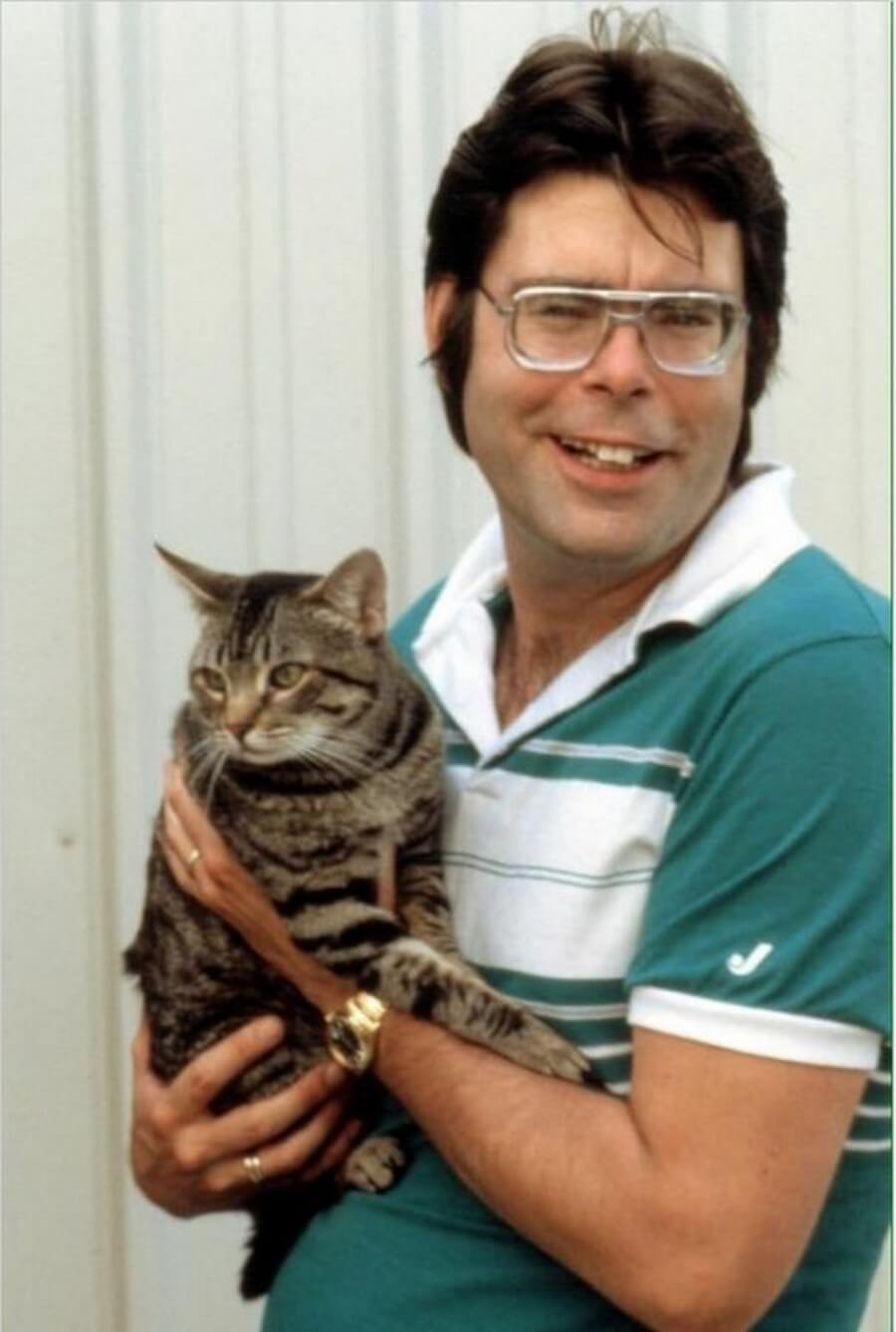 Photograph of horror author Stephen King wearing glasses and a green and white striped shirt, holding a tabby cat.