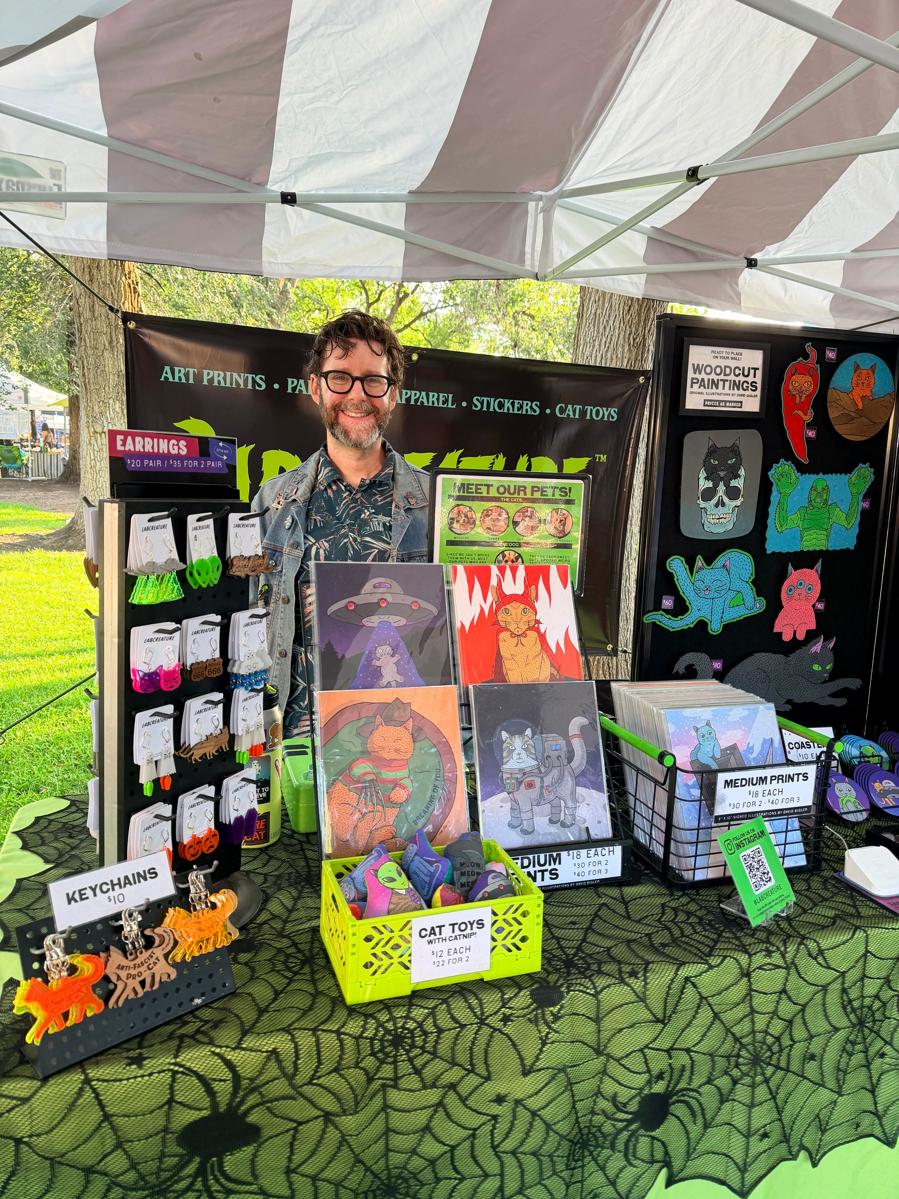 Photo of artist David Bigler standing behind a table at an outdoor market booth displaying various colorful cat-themed art prints, keychains, earrings, and cat toys.