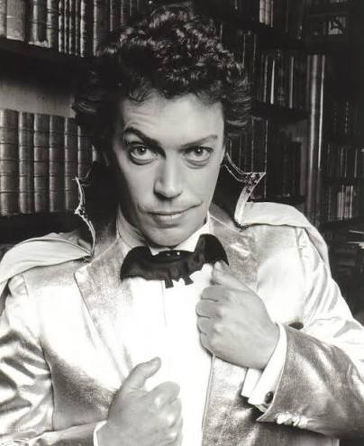 Black and white photo of actor Tim Curry from 1986’s The Worst Witch dressed in a shiny suit with a bat bow tie, posing in front of a bookshelf filled with books with a wry smile and upturned right eyebrow. He has both hands on the front of his jacket. 